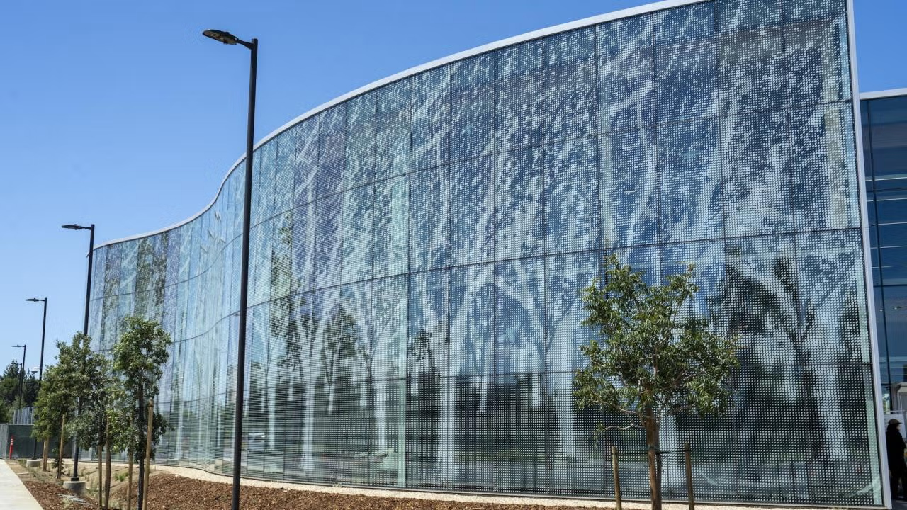Curved glass facade with white tree silhouettes and young trees along sidewalk