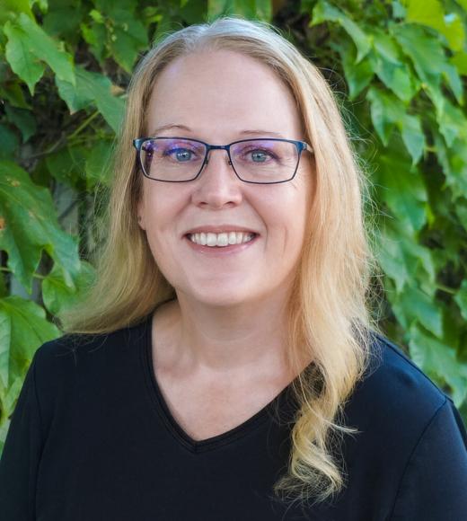 Smiling headshot of woman with glasses, long blonde hair, black top, leafy background