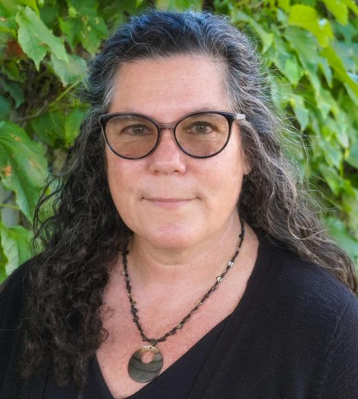 Headshot of a middle-aged woman with gray-streaked curly hair, glasses, black top, vine backdrop