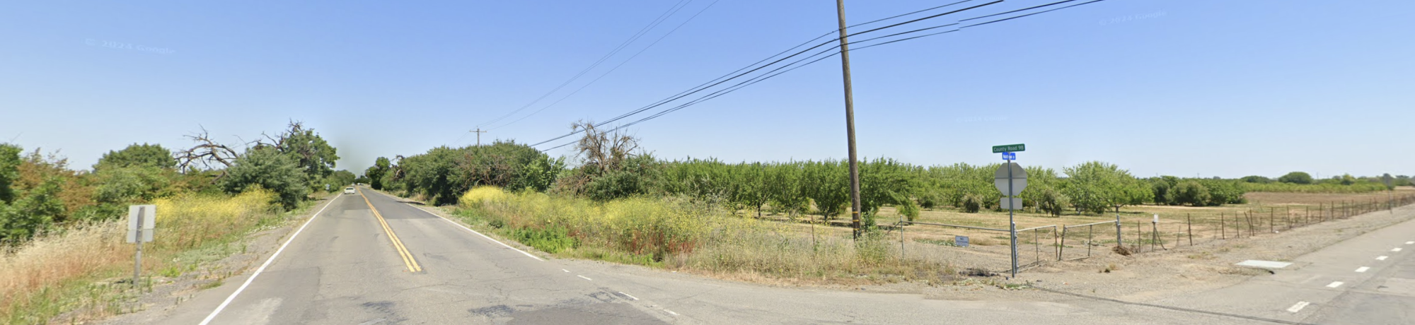 Rural two-lane road intersection with dry fields and power lines under a blue sky.